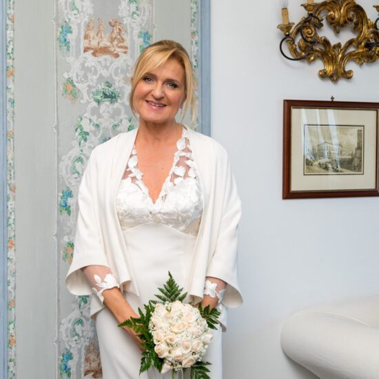 Matrimoni, sposa in piedi davanti un muro affrescato con il bouquet fra le mani, sguardo dolce in macchina