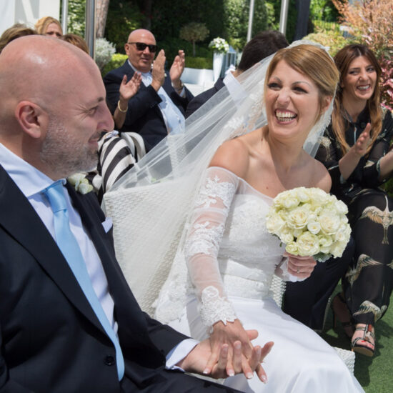 Matrimoni, sposa con bouquet seduta che ride guardando il marito che sorride, dietro gli invitati applaudono
