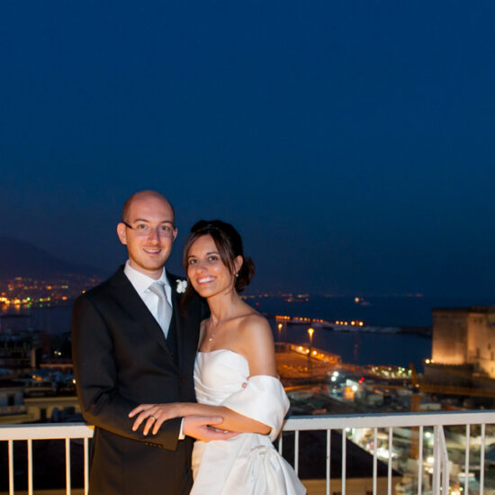 Matrimoni, sposi abbracciati che guardano verso il fotografo, sfondo notturno con maschio angioino mare e vesuvio, predominanza blu per lo sfondo e gialla-arancio per le parti illuminate