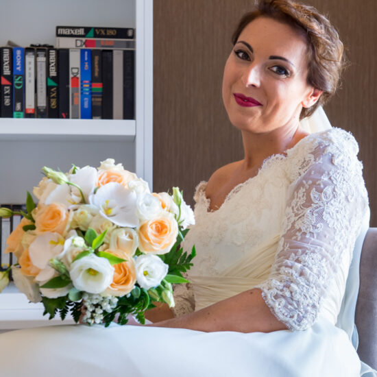 Matrimoni, sposa seduta, spalle alla finestra con bouquet sferico in mano con rose bianche e arancioni, sguardo dolce in camera