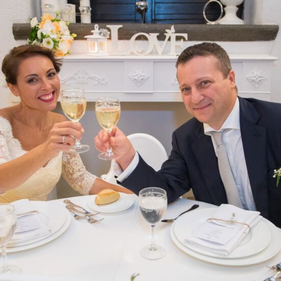 Matrimoni, gli sposi avvicinano i calici guardando il fotografo mentre sono seduti a tavola prima dei pasti all'inizio del ricevimento