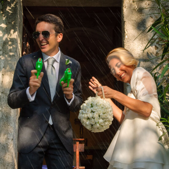Matrimoni, sposi all'uscita della chiesa sono accolti da una intensa pioggia di riso, lui si protegge con occhiali da sole e pistole ad acqua, lei indietreggia per proteggersi
