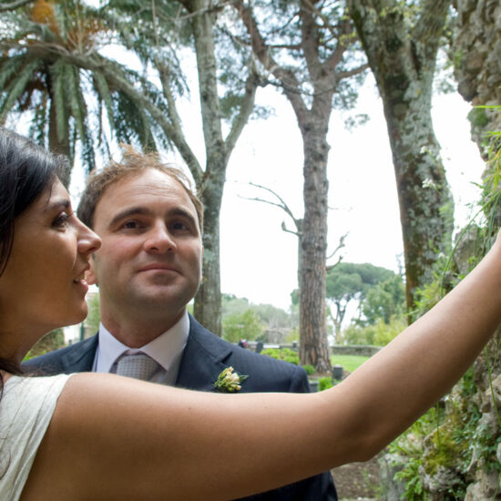 Matrimoni, sposi in giardino, lei prende un fiorellino da un muro di pietra, lui guarda me e dietro degli alberi alti