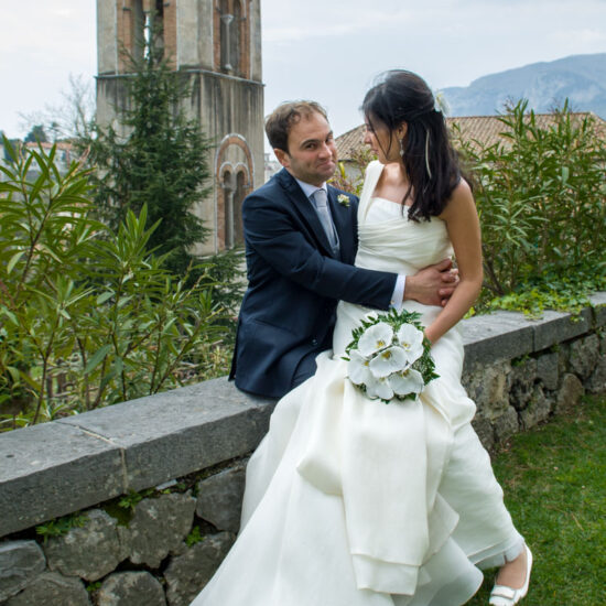 Matrimoni, sposi seduti sul muretto di un giardino, campanile alle spalle, lui guarda me con viso scherzoso e lei guarda lui