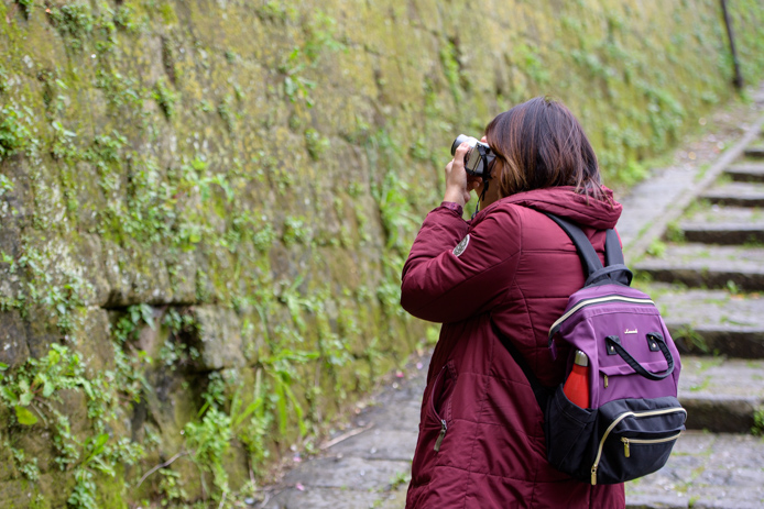 modulo base del corso di fotografia ragazza con giubbino bordeaux che scatta qualcosa su di una parete di tufo ricoperta di vegetazione