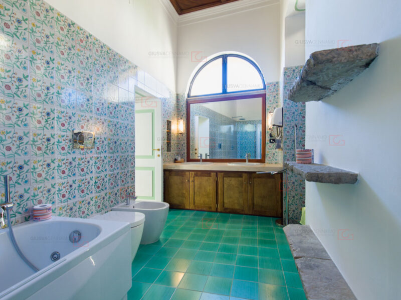 fotografare gli interni - real estate, bagno con maioliche verdi, grande specchi e due lavandini. Vasca e igienici, mensole in pietra, finestrino a semicerchio.