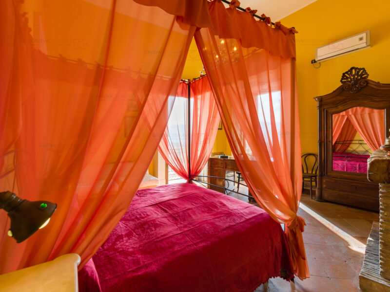 fotografare gli interni - real estate, letto con copriletto rosso con baldacchino e tendaggi arancioni, armadio in legno con specchiera sull'anta frontale, due balconi s'intravedono attraverso i drappeggi.