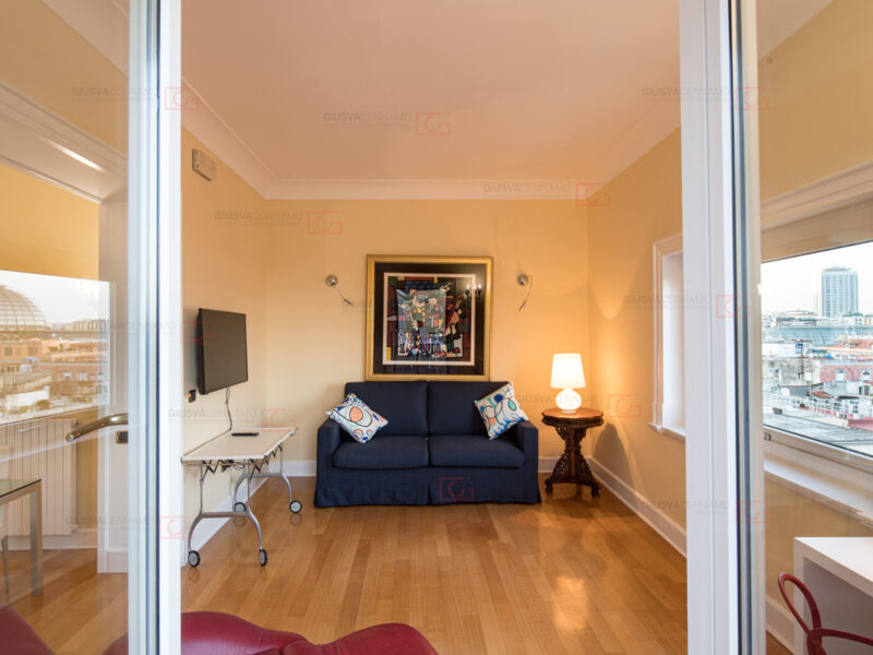 fotografare gli interni - real estate, vista dal balcone verso la stanza dalle pareti gialle, divano blu in fondo e finestra sulla destra. Riflessi sulle ante delle porte del balcone in particolare si vede la cupola della galleria vittoria. Quadro stile matisse sopra il divano.