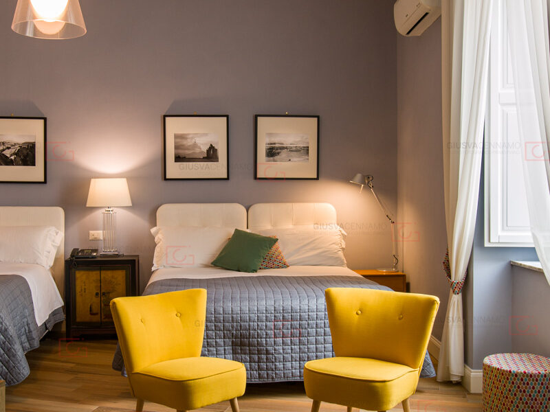 fotografare gli interni - real estate, stanza dalle pareti grigio chiaro, finestra a destra e due poltroncine gialle ai piedi di un letto frontale