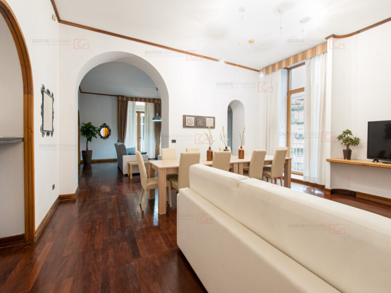 fotografare gli interni - real estate, ampio salone divano bejge, tavolo in legno chiaro con 10 sedie, arco comunicante con secondo salone con divano specchio e pianta. balconi in entrambi