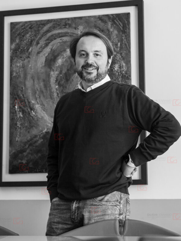 Ritratto a un professionista, CEO in azienda software in felpa nera su camicia bianca. Braccio sinistro piegato con mano sul fianco. In piedi, sorridente, con sguardo in camera. Sfondo con quadro che rende un movimento circolare.