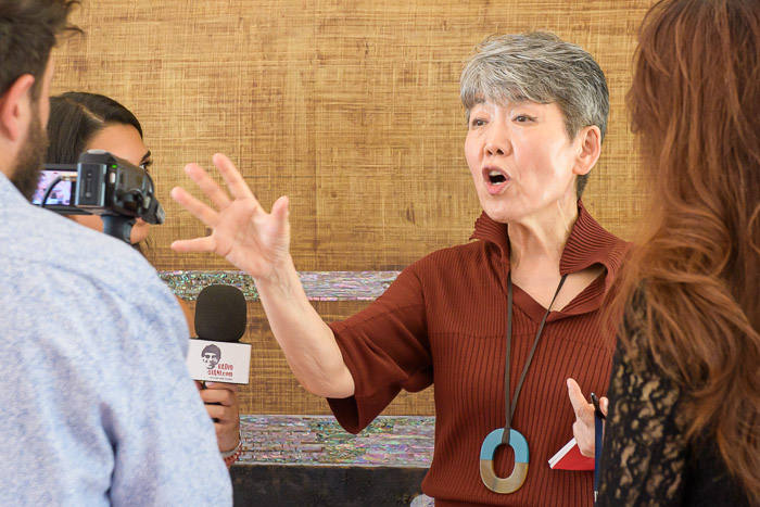 Giusva servizi fotografici - ritratti ad eventi - artista donna coreana che spiega la sua opera gesticolando con un giornalista sullo sfondo di un suo quadro