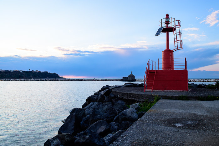 Giusva servizi fotografici - Procida paesaggi, sul molo, faro rosso spento sulla destra alle ultime luci del crepuscolo, a sinistra il mare, un'altro molo in fondo e poi la collina. Aldi sopra cielo azzurro con nuvole nella parte bassa