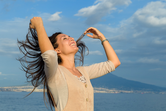 Giusva servizi fotografici - Ritratti privati, donna girata a tre quarti con un bel sorriso, le mani che muovono i capelli, luce calda del tramonto su di lei e sfondo azzurro cielo e parte del vesuvio